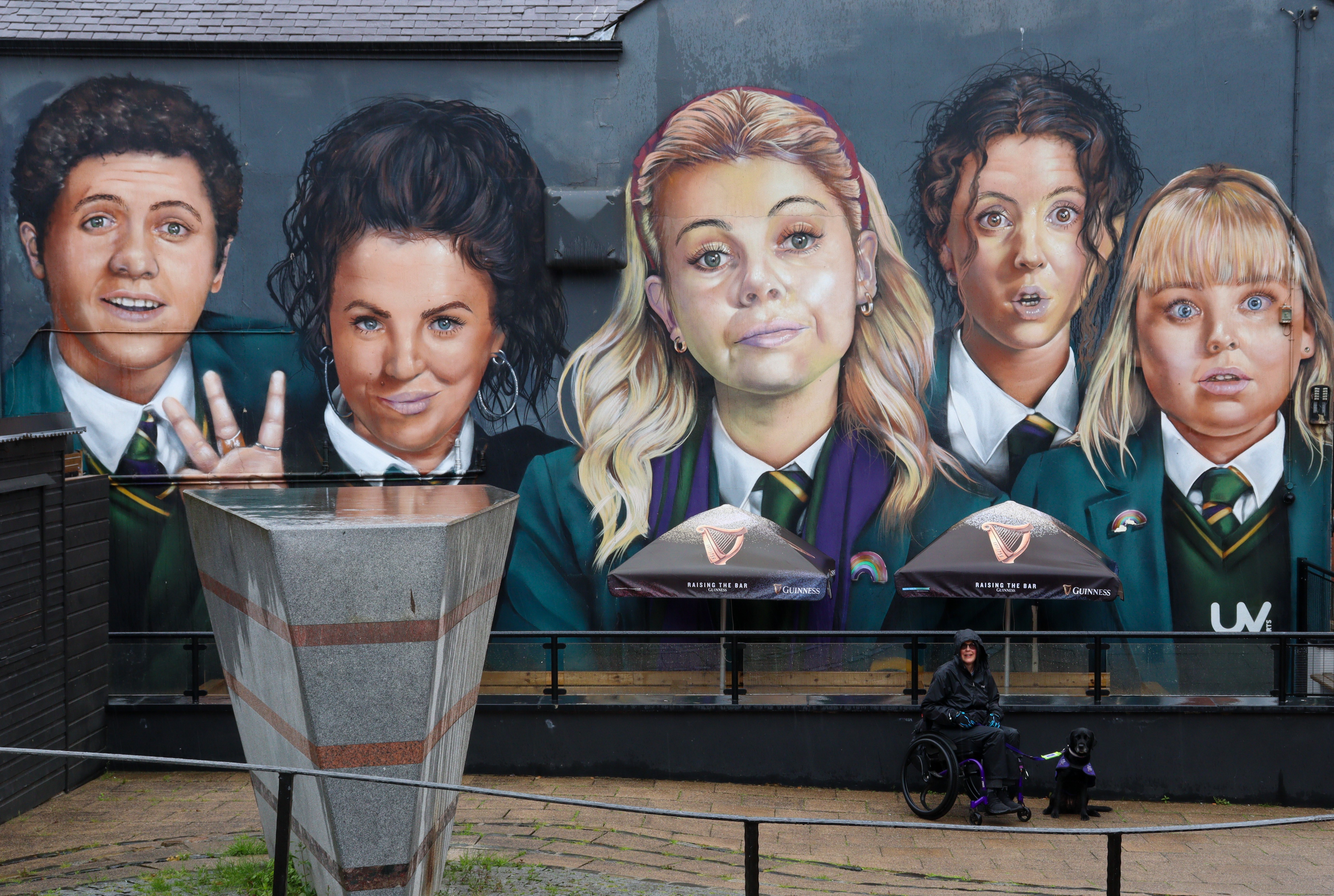 Large mural of four girls and one boy in school uniform. A lady in a wheelchair with a dog is sitting in front of it.