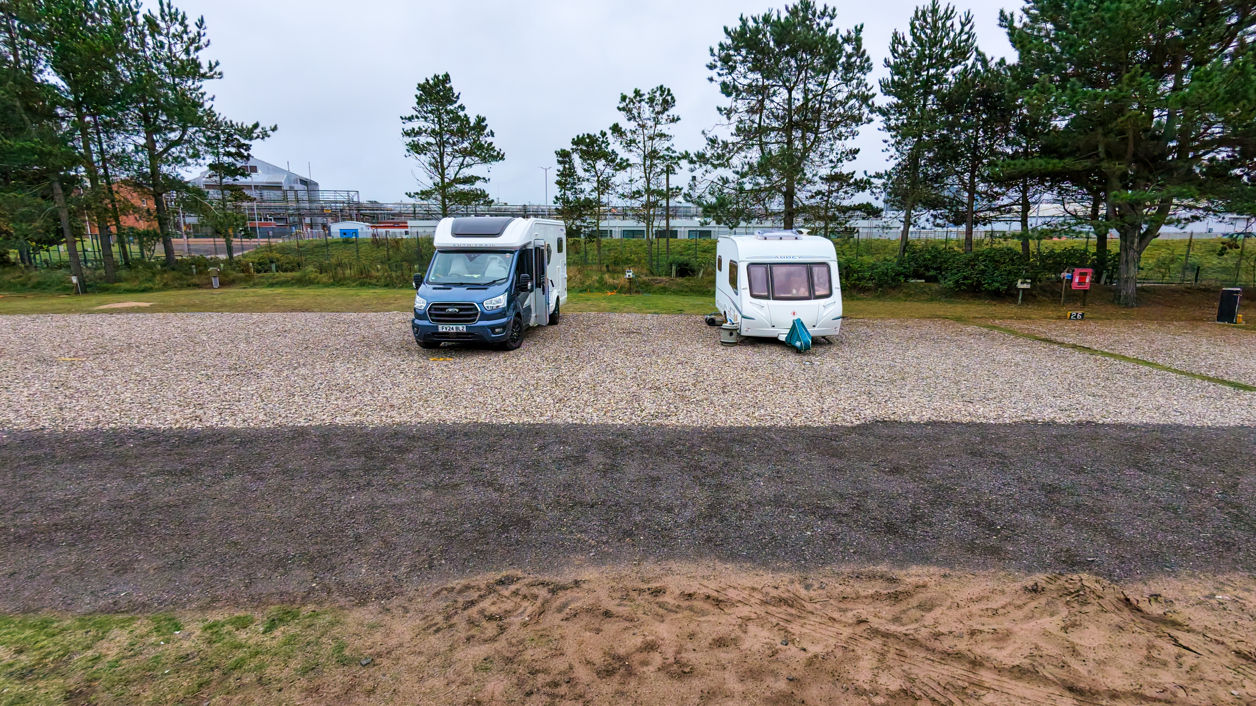 A motorhome and a caravan parked quite close together on a pebble surface with trees behind.