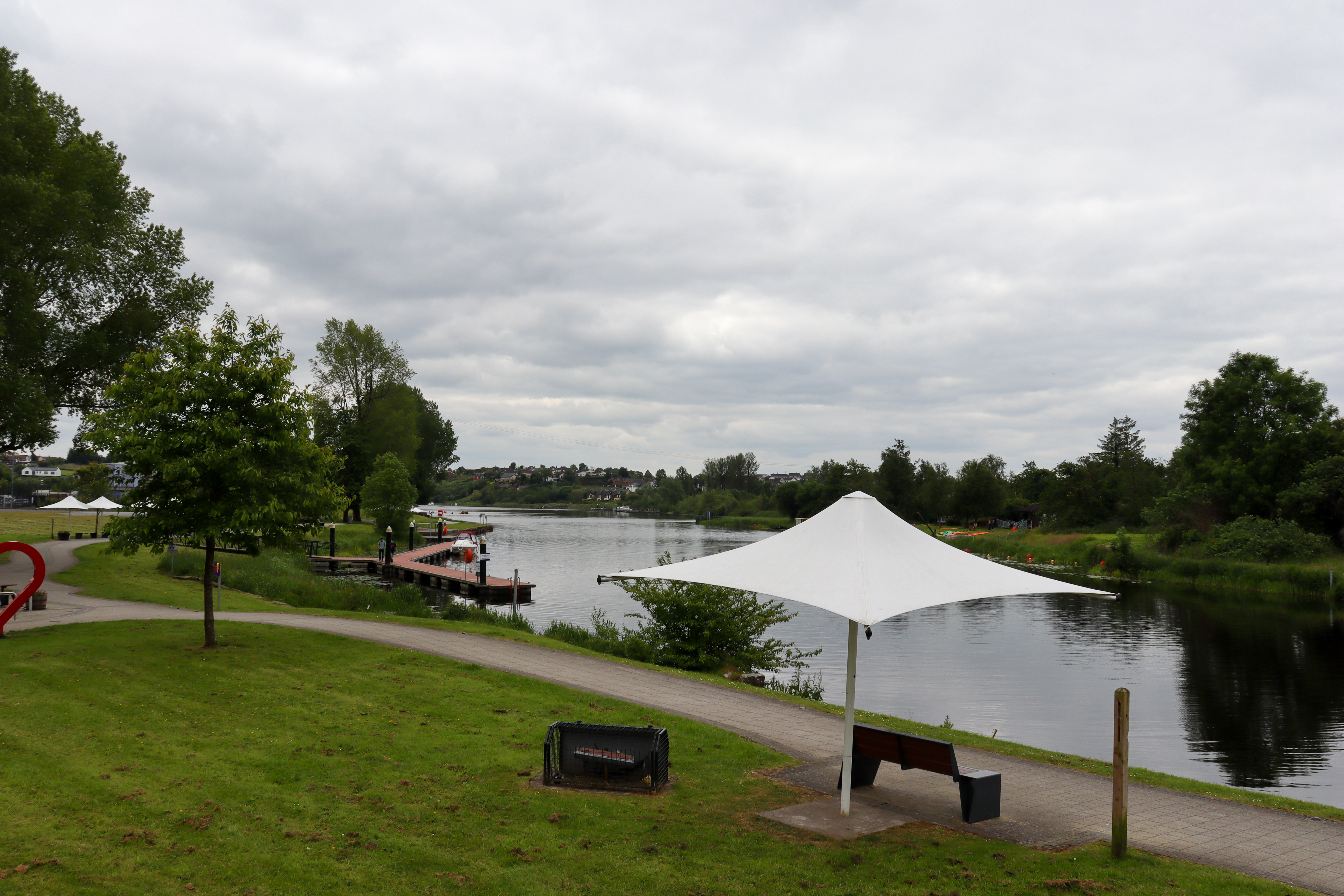 Landscaped gardens with wide path next to the water, with a bridge in the distance.