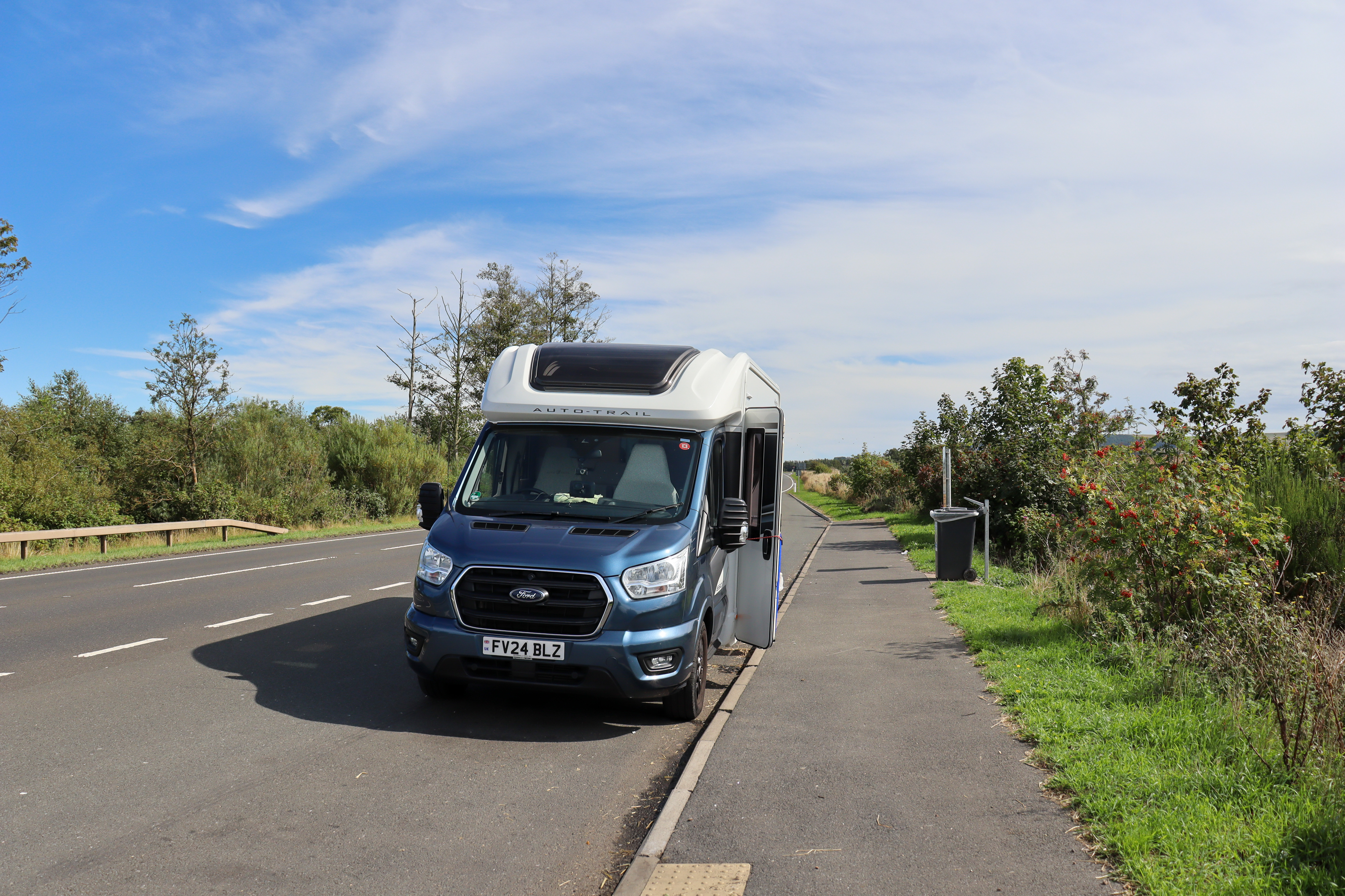 Motorhome parked in a layby with a wide pavement and large bin at the side.