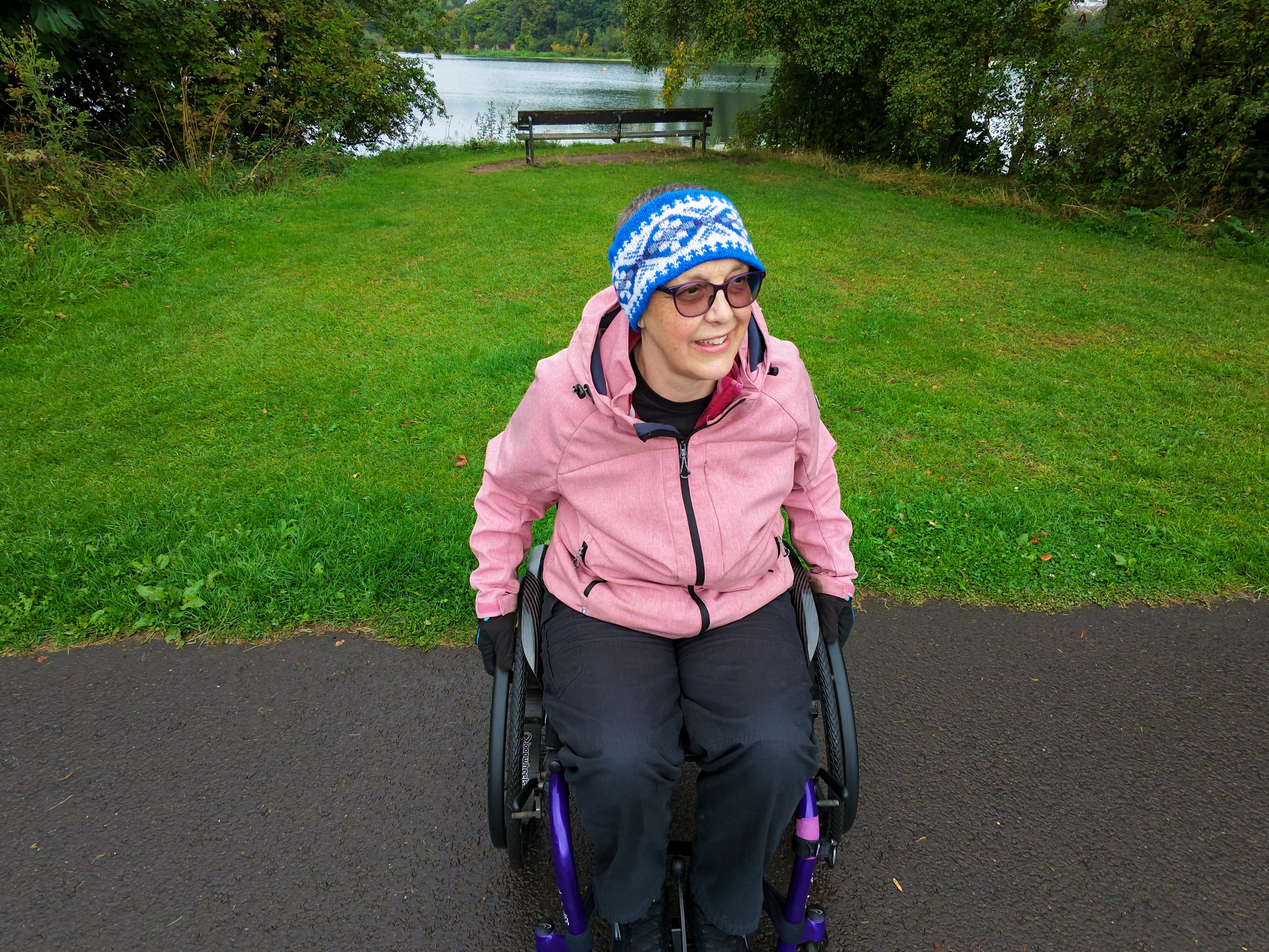 Lady in a pink coat and blue headband, sitting in a wheelchair in front of a loch surrounded by trees.