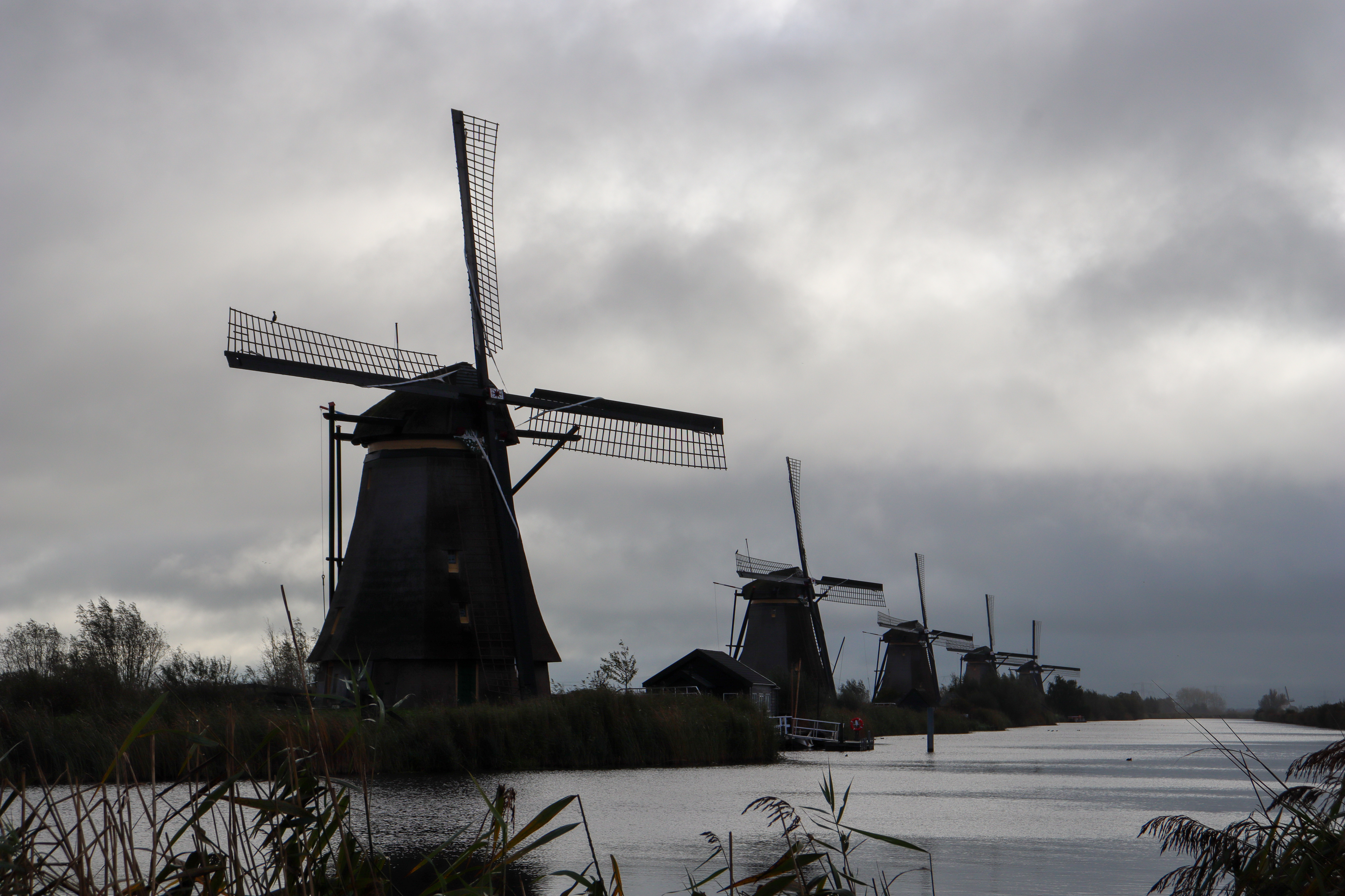 5 windmills along a river bank with very cloudy skies