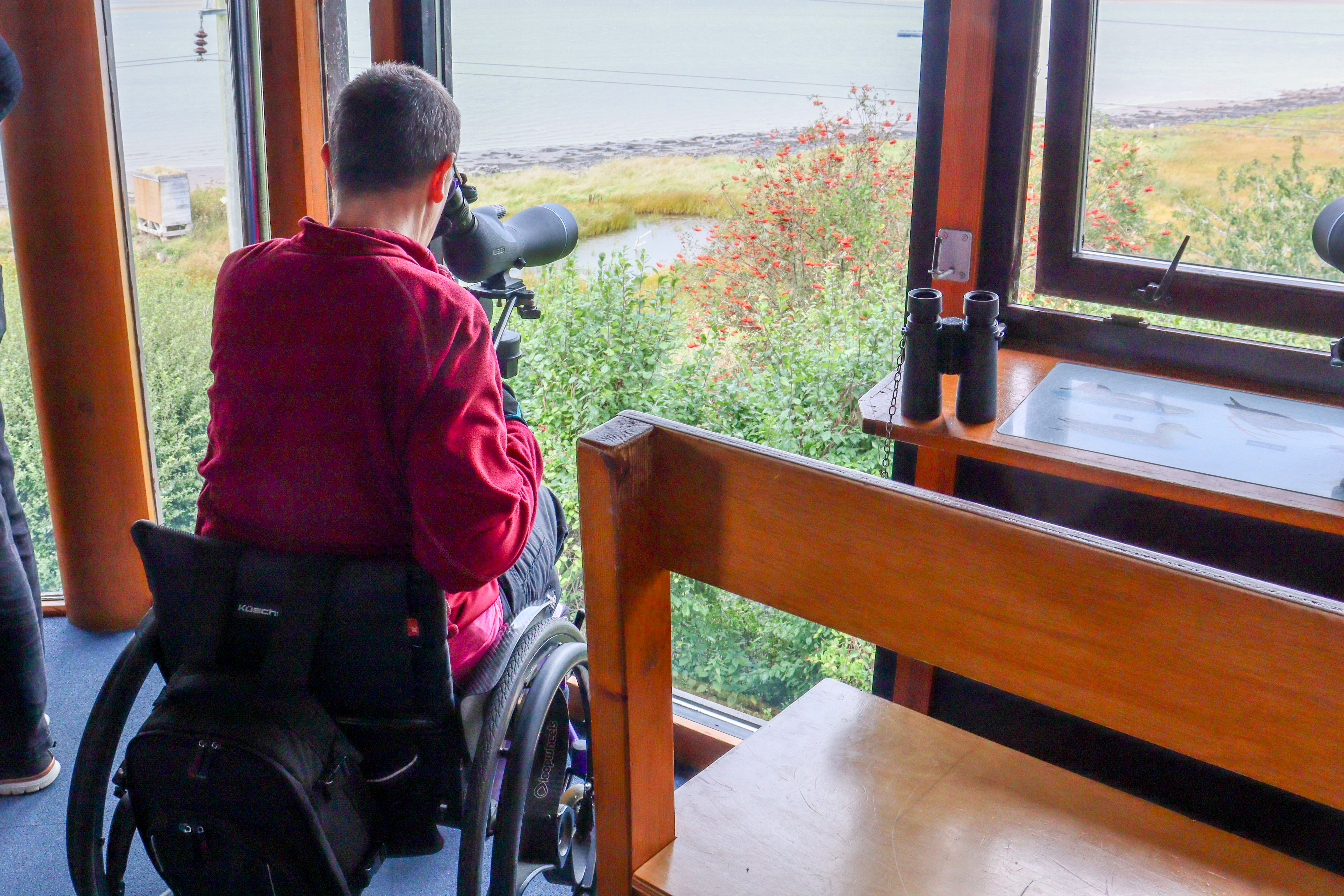 Lady in a wheelchair looking out of a full length window with a telescope