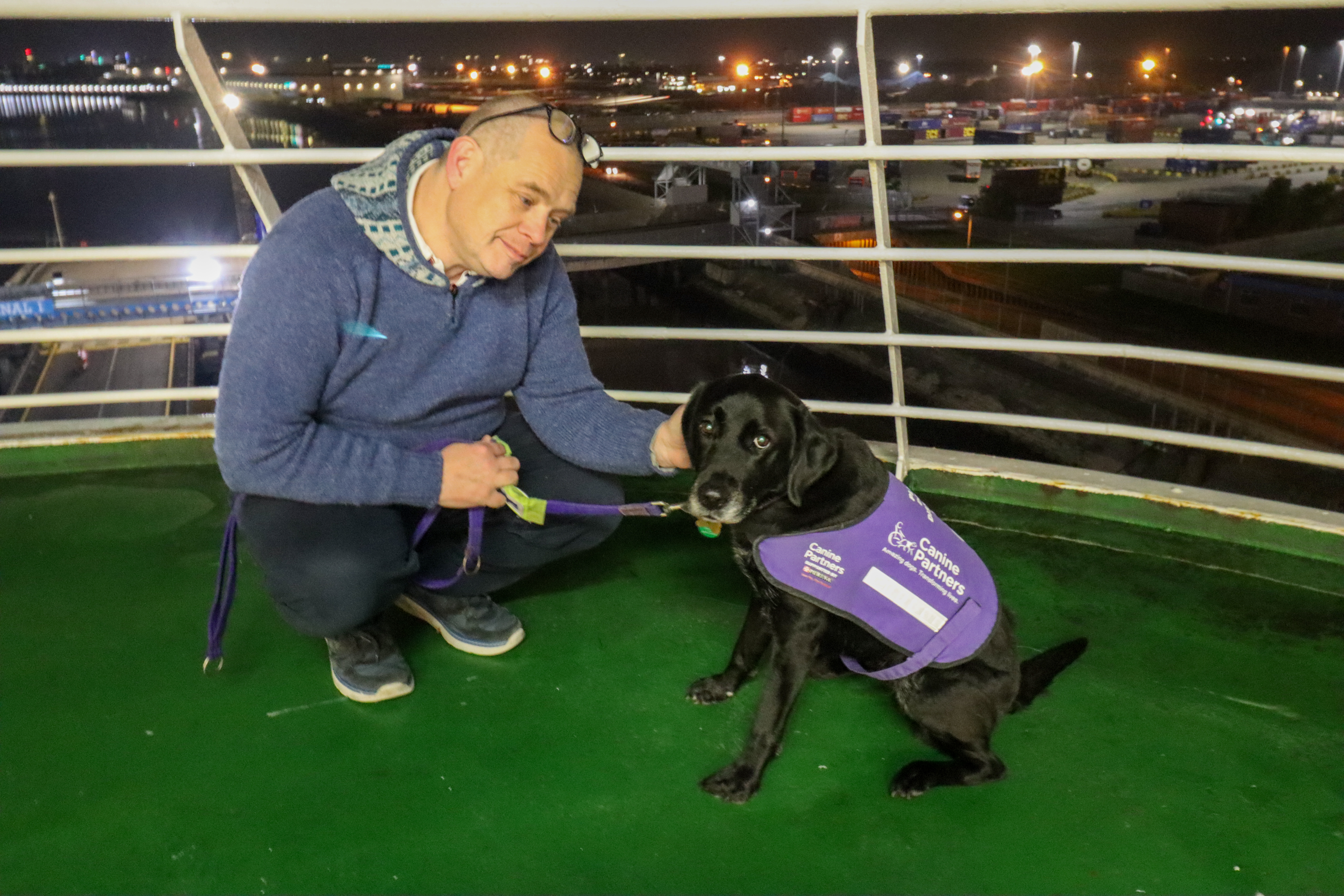 Man with a black labrador wearing a purple Canine Partners jacket, on an outdoor deck with railings behind.