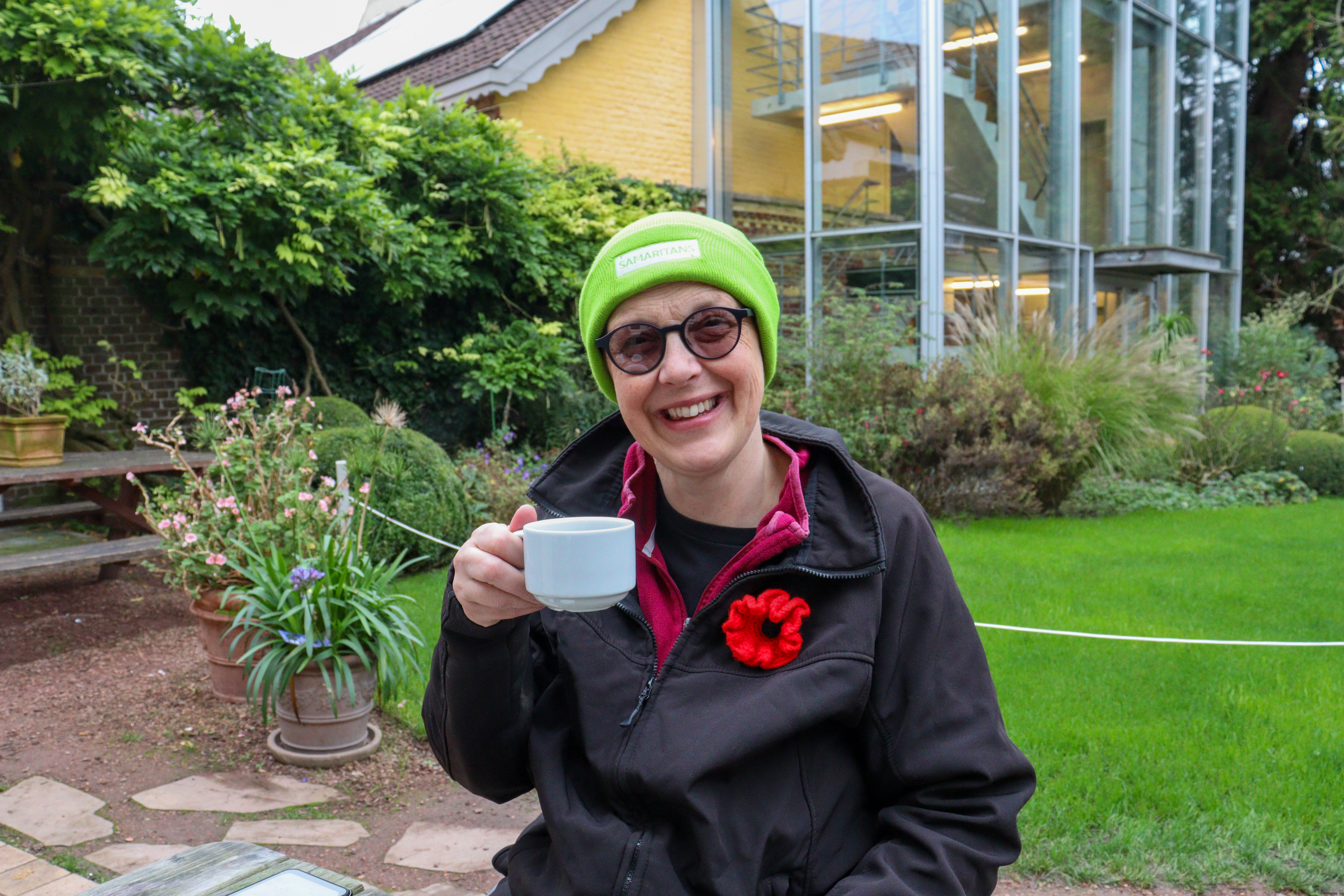 Lady in a hat and coat, wearing a woolly red poppy, holding a cup of coffee