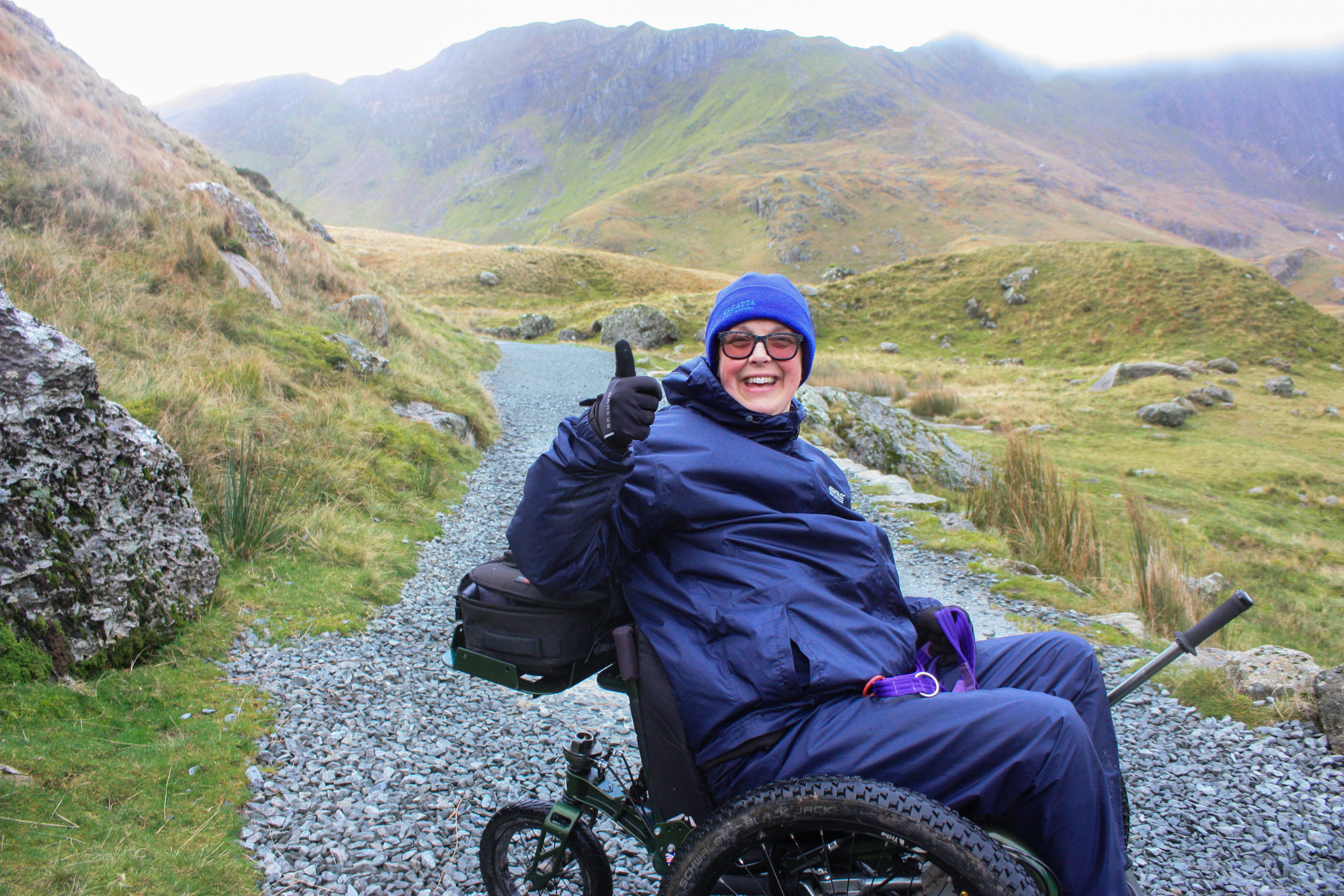Lady in full waterproofs, sitting in her mountain trike on a mountain path, with a huge smile and thumbs up.