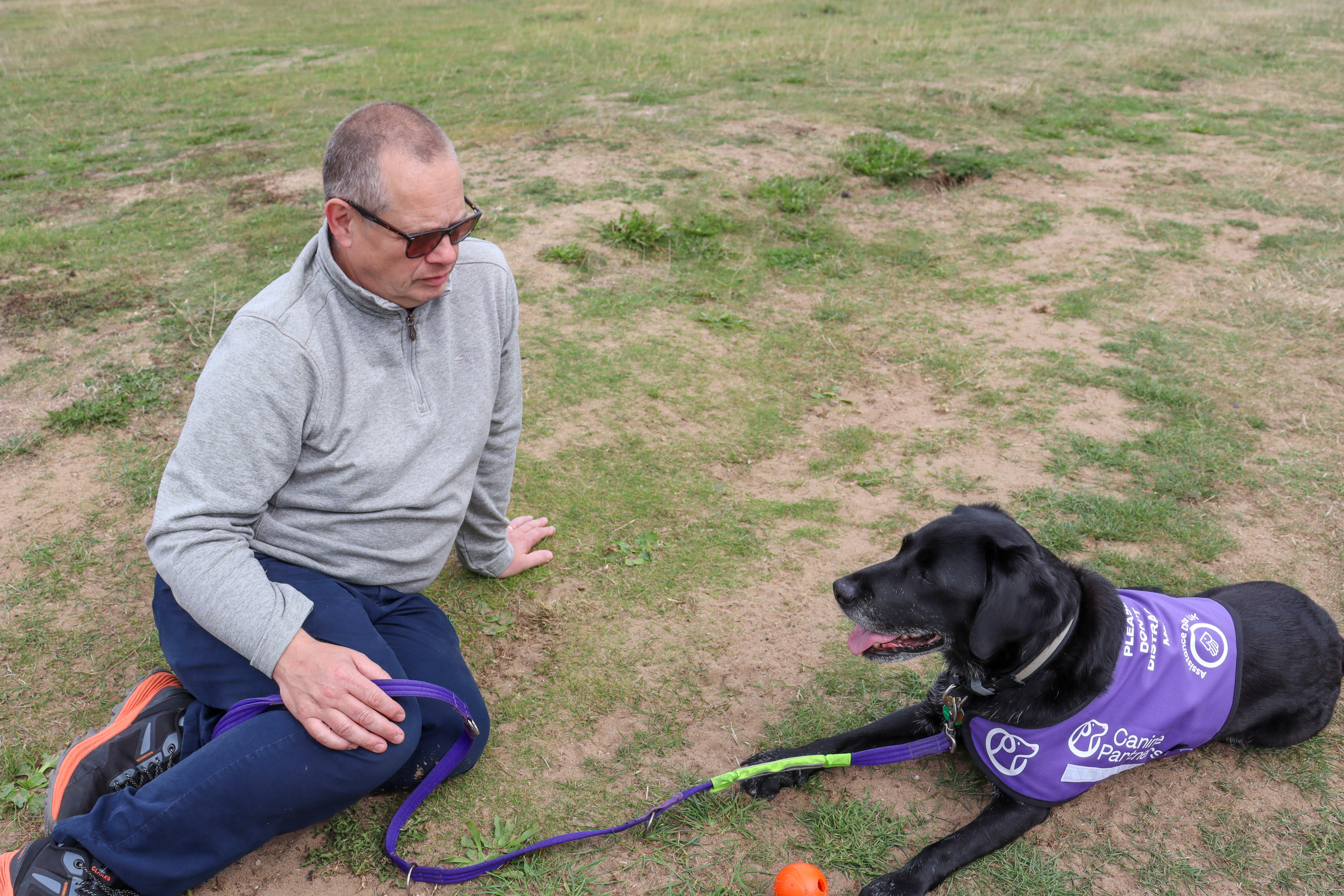 A man and a black Labrador sitting together on the grass, looking into each other's eyes.