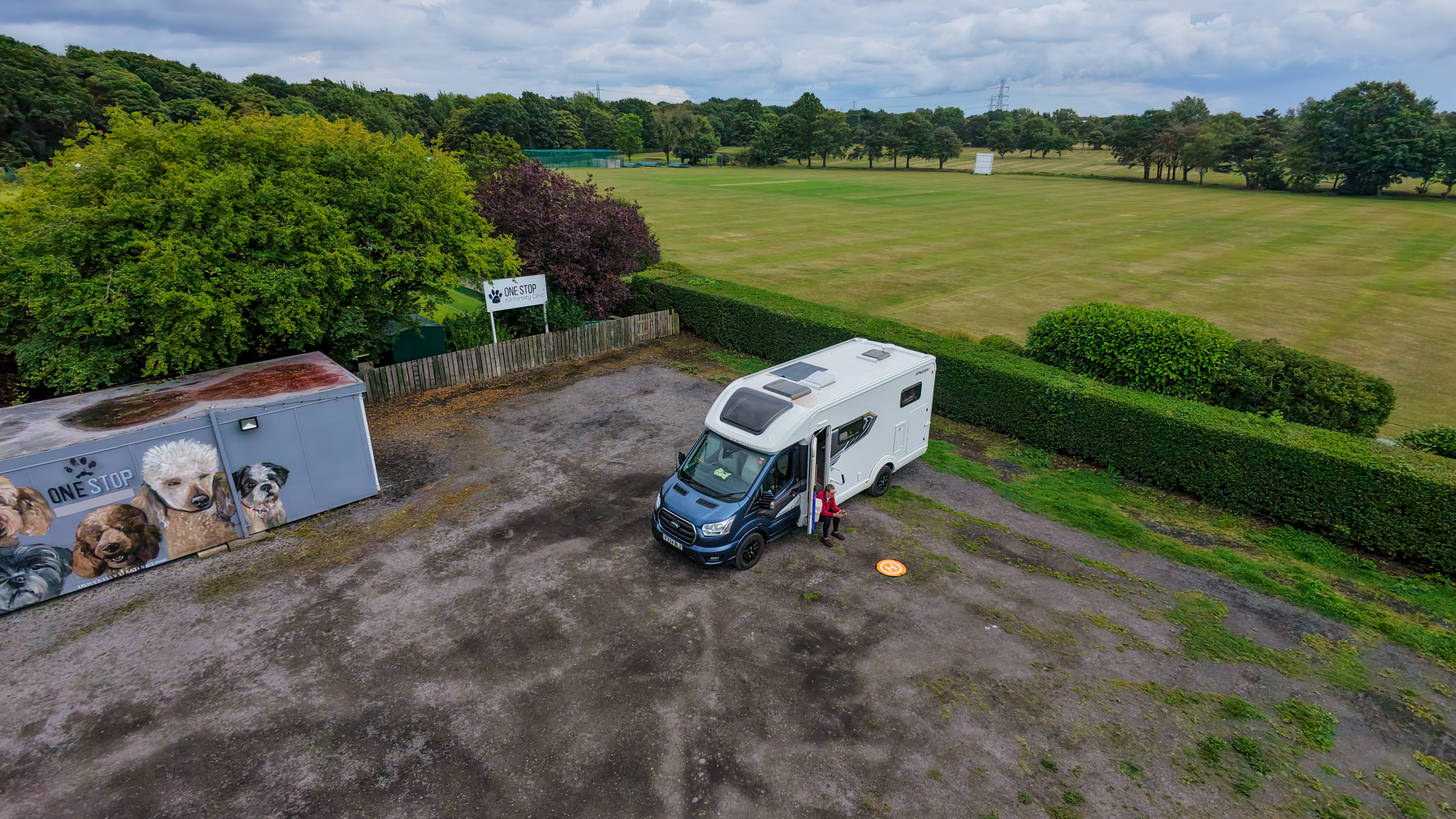 Aerial view of a motorhome with a portacabin in the same car park and a cricket pitch behind.