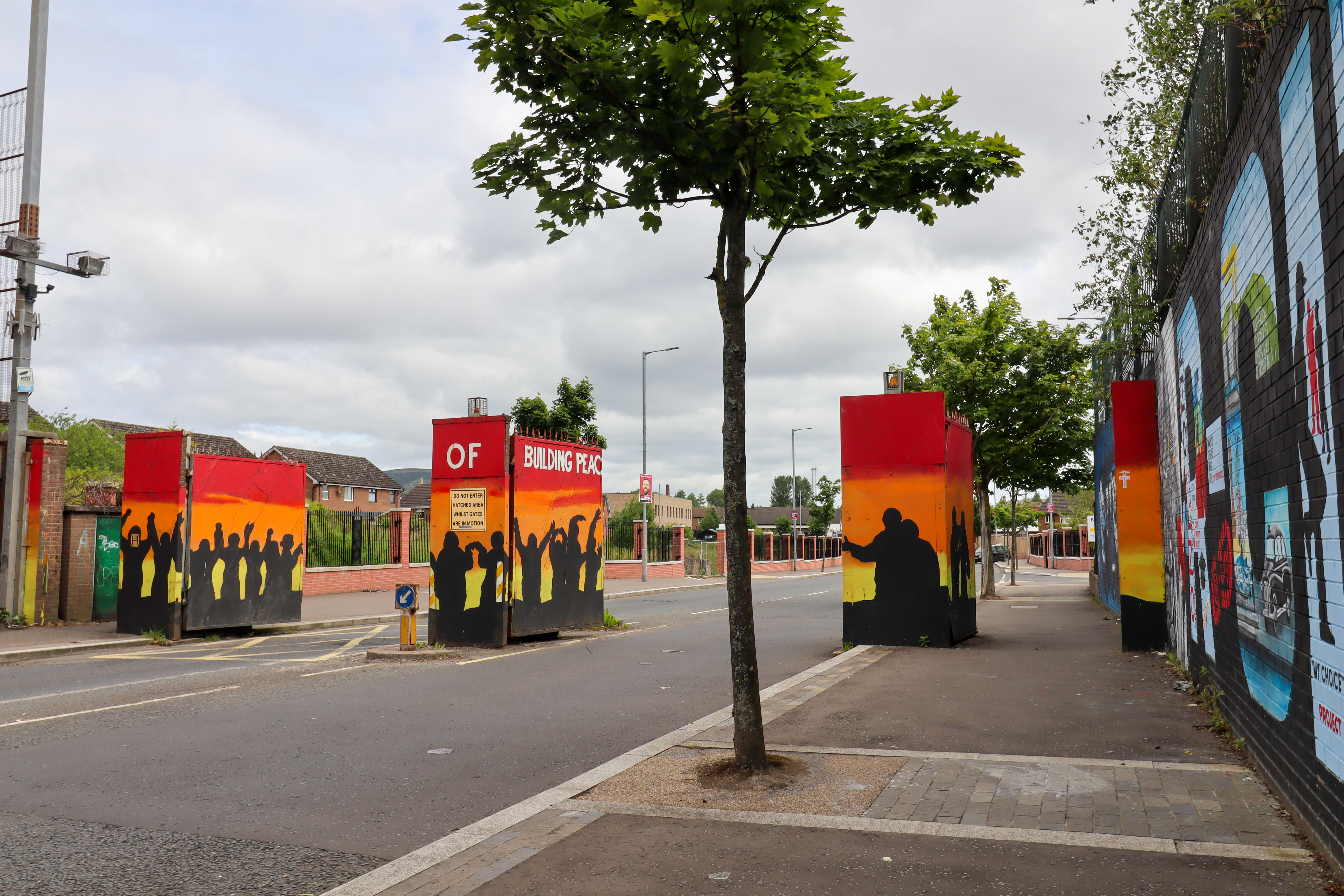 Large security gates covered in paintings about peace, with the peacewall going down the road on the right and a normal housing estate on the left.