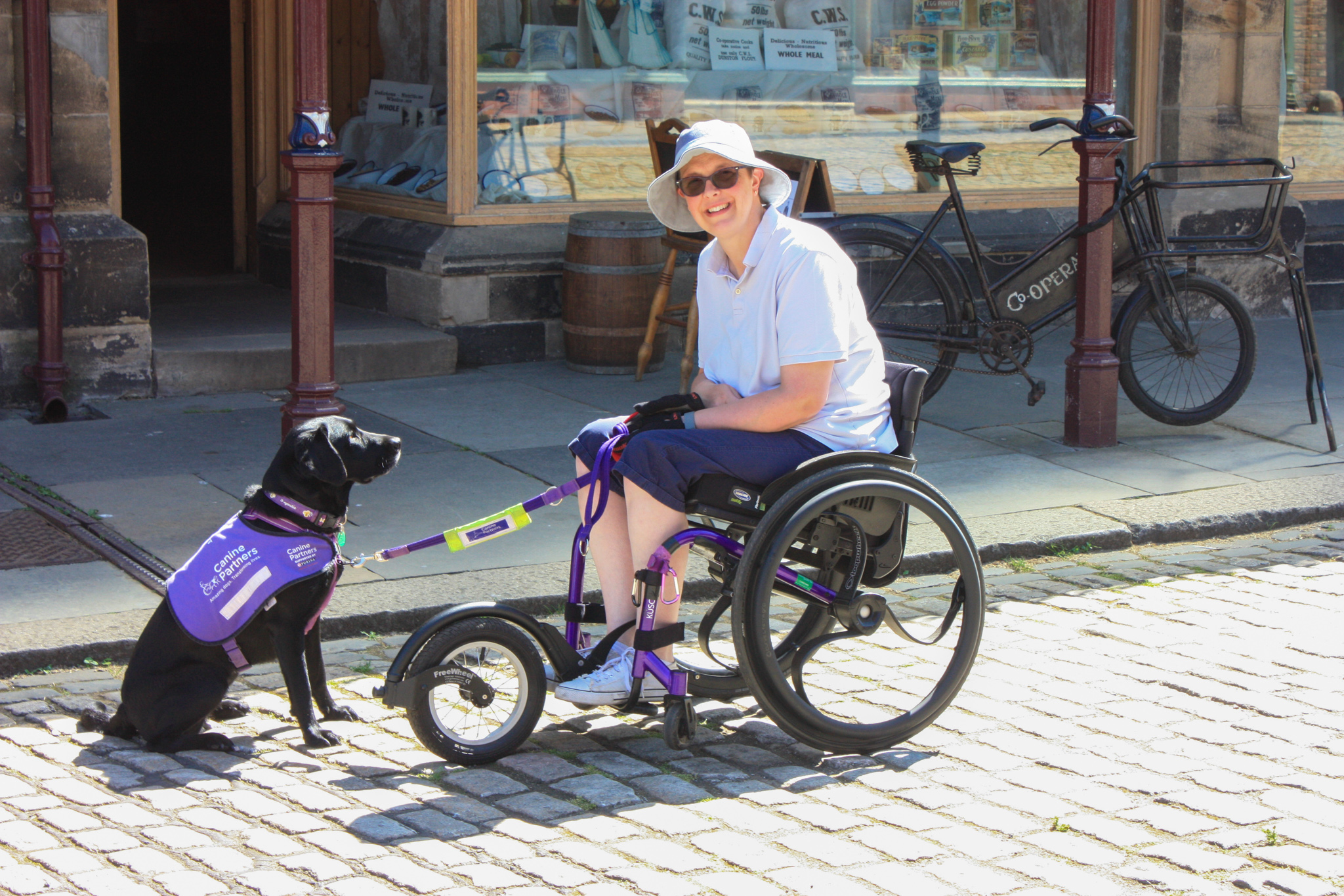 Nicki in a manual wheelchair with additional big front wheel attachment. Canine Partner Liggy is sitting in front of her on a cobbled street.