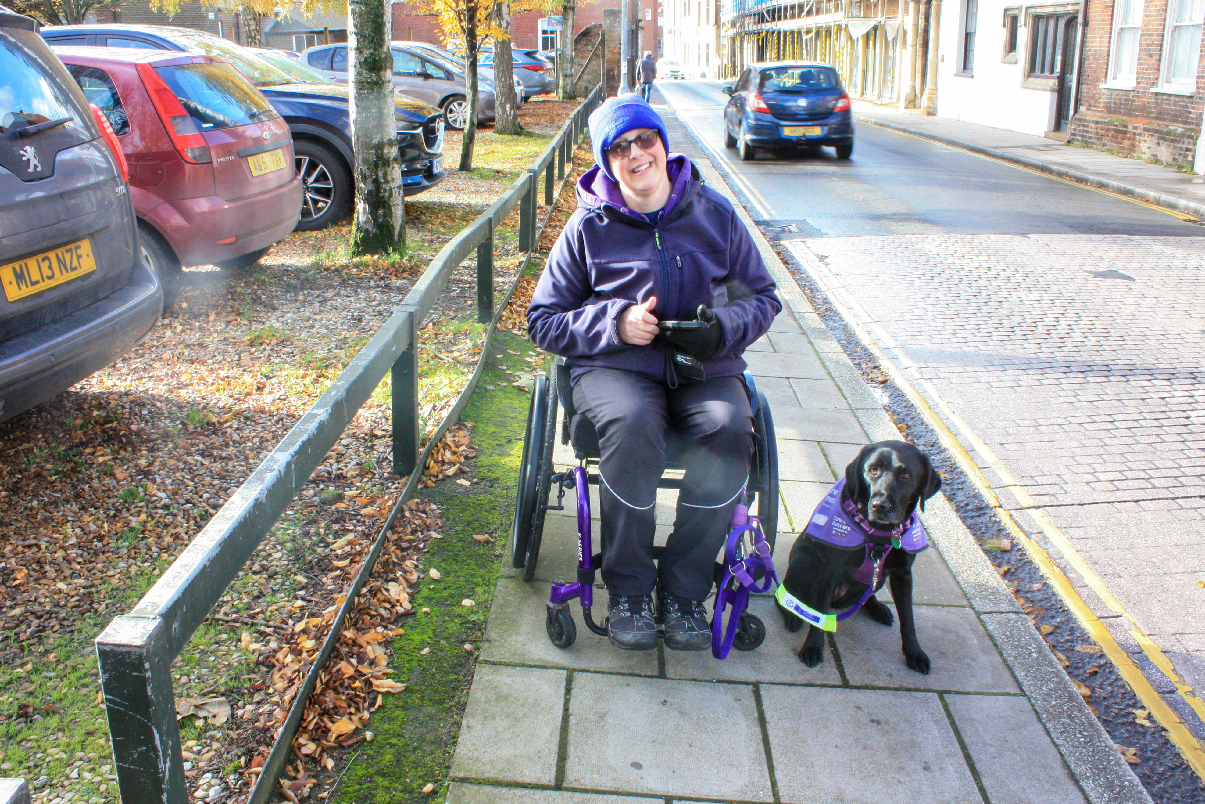 Nicki in a manual wheelchair with Canine Partner Liggy sitting next to her. They are on a normal town pavement.
